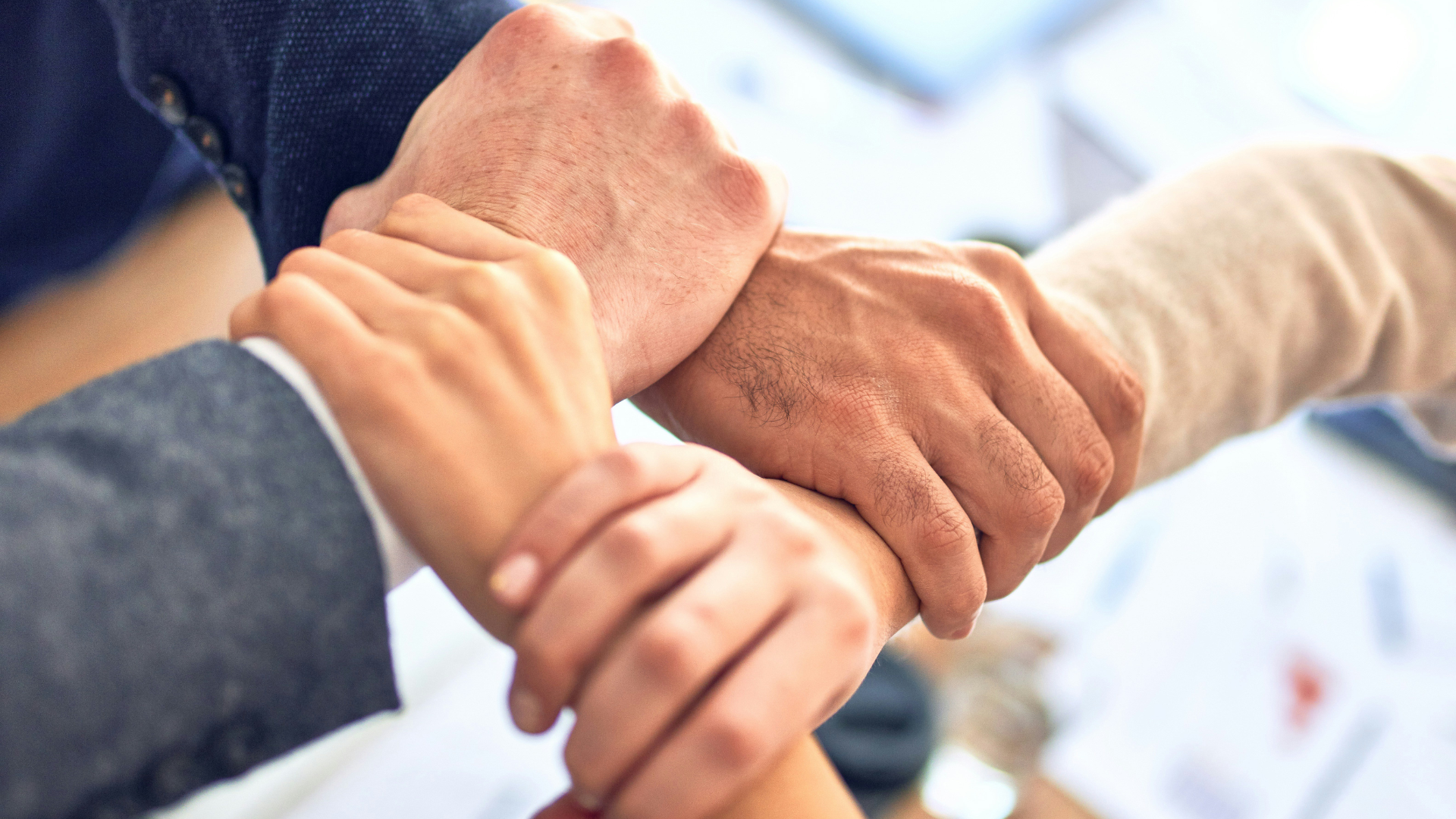 Zespół osób w biurze składa dłonie jedna na drugiej na środku stołu, symbol współpracy i wspólnego celu.